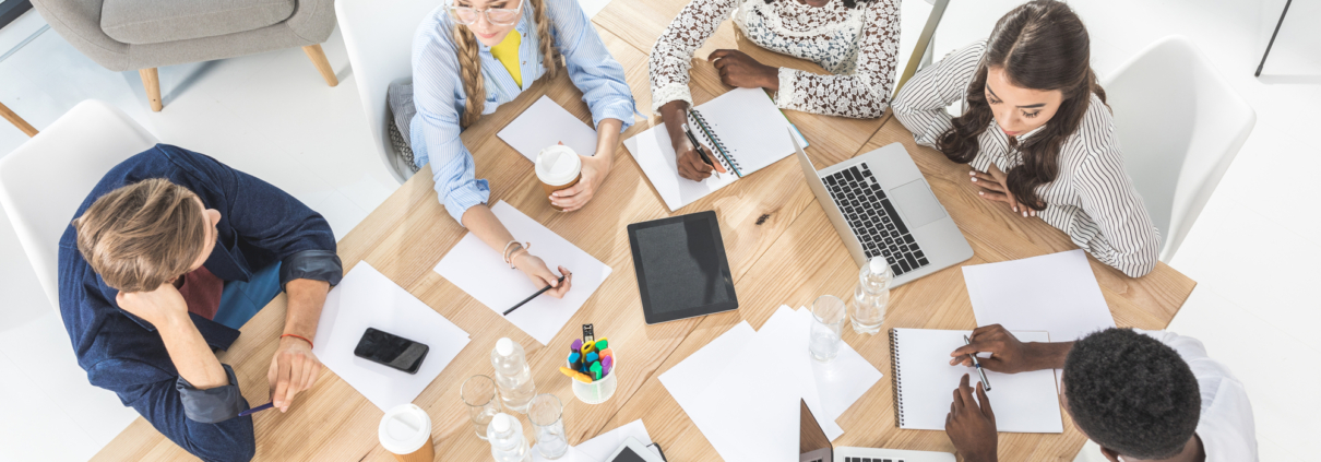 Eine Gruppe von Menschen sitzt an einem Tisch, arbeitet zusammen und macht Notizen mit Laptops, Tablets und Schreibmaterial.