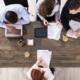 Vier Personen arbeiten an einem Tisch mit Laptops und Dokumenten. Kaffeetassen und Schreibutensilien sind verteilt.