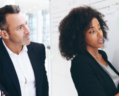 Drei Personen besprechen und analysieren Informationen, die auf einem Whiteboard stehen. Eine Frau erklärt dabei den Inhalt.