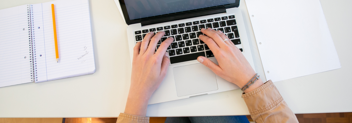 Eine Person arbeitet an einem Laptop mit Notizbüchern und Papieren auf dem Schreibtisch.