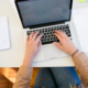 Eine Person arbeitet an einem Laptop mit Notizbüchern und Papieren auf dem Schreibtisch.