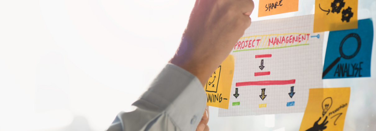 Hand, die eine Markierung an einem Whiteboard mit bunten Notizzetteln und einem Projektplan vornimmt.
