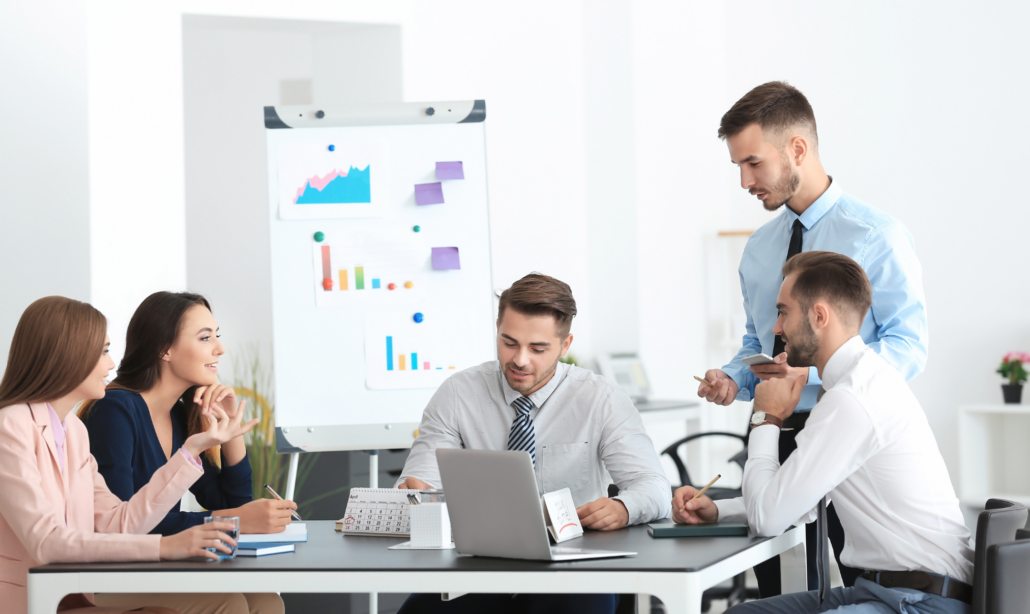 Fünf Personen in einem Meeting, sitzend um einen Tisch, mit einem Laptop und einer Flipchart mit Diagrammen im Hintergrund.