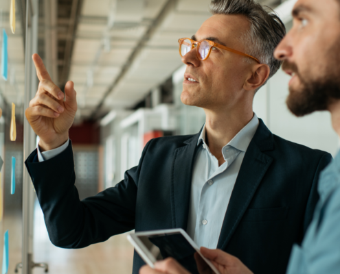 Zwei Männer diskutieren und zeigen auf Notizen, die an einer Glaswand angebracht sind. Einer der Männer hält ein Tablet in der Hand.