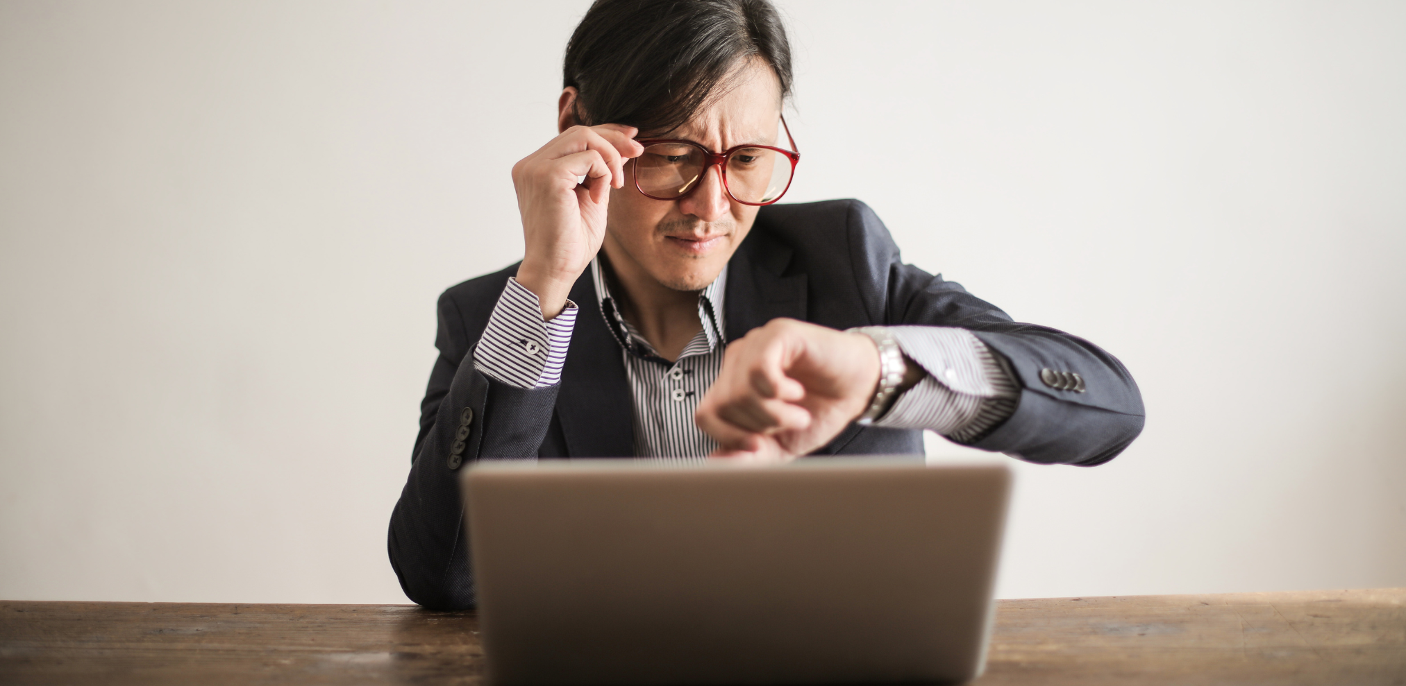 Person in Anzug blickt auf ihre Armbanduhr, während sie vor einem Laptop sitzt.