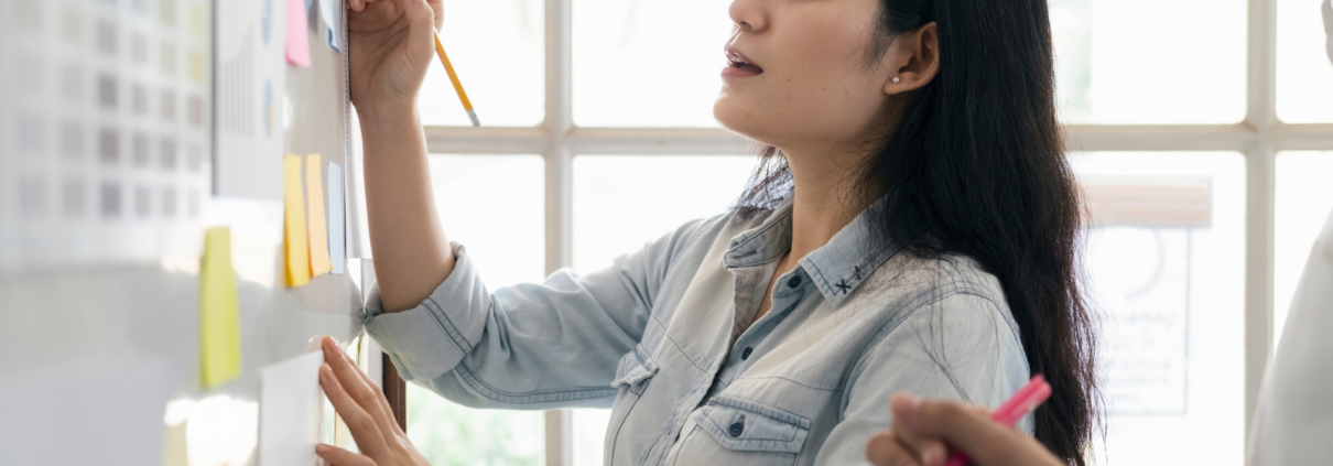 Eine Frau schreibt mit einem Bleistift auf eine Tafel, die mit verschiedenen farbigen Haftnotizen und Diagrammen bedeckt ist. Im Hintergrund befindet sich ein großes Fenster, das viel Tageslicht hereinlässt. Eine andere Person hält ein Notizbuch und einen Stift, als ob sie sich Notizen macht.