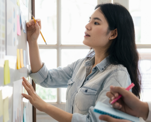 Eine Frau schreibt mit einem Bleistift auf eine Tafel, die mit verschiedenen farbigen Haftnotizen und Diagrammen bedeckt ist. Im Hintergrund befindet sich ein großes Fenster, das viel Tageslicht hereinlässt. Eine andere Person hält ein Notizbuch und einen Stift, als ob sie sich Notizen macht.