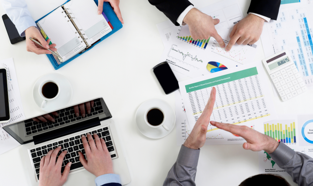 Das Bild zeigt ein Team bei einer Besprechung, wobei verschiedene Personen Dokumente mit Diagrammen und Statistiken überprüfen und besprechen. Auf dem Tisch befinden sich ein Laptop, Kaffeetassen und weitere Arbeitsmaterialien.