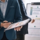 Geschäftspersonen besprechen Diagramme auf einem Clipboard, während andere auf eine Tafel mit Diagrammen und Notizen im Hintergrund zeigen.
