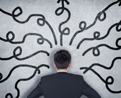 Rückenansicht eines Mannes in einem Anzug, der vor einer grauen Wand steht, auf der viele schwarze, verschlungene Pfeile in verschiedene Richtungen zeigen.