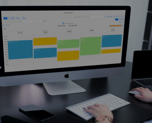 Eine Person arbeitet an einem Schreibtisch mit einem großen iMac, auf dessen Bildschirm eine TimeTrack Wochenansicht eines Kalenders mit farblich markierten Terminen angezeigt wird.