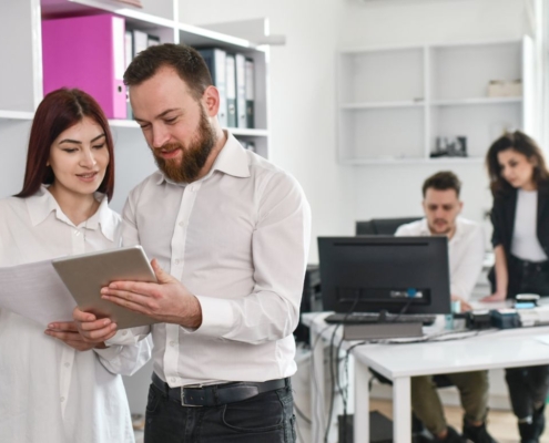 Zwei Kollegen in einem Büro betrachten gemeinsam ein Tablet, während im Hintergrund zwei weitere Personen an einem Schreibtisch arbeiten.
