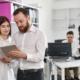 Zwei Kollegen in einem Büro betrachten gemeinsam ein Tablet, während im Hintergrund zwei weitere Personen an einem Schreibtisch arbeiten.