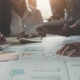 Mehrere Personen arbeiten an einem Tisch, der mit Finanzberichten und Diagrammen bedeckt ist, während sie mit einem Tablet und einem Taschenrechner arbeiten.