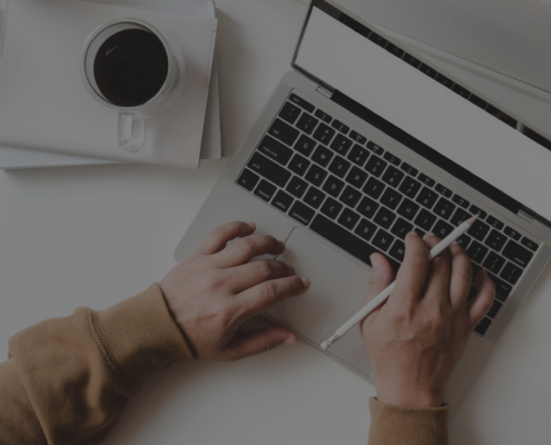 Das Bild zeigt eine Person, die an einem Laptop arbeitet. Auf dem Schreibtisch befinden sich eine Tasse Kaffee, ein Notizbuch, Bleistifte und ein Stifthalter. Die Person hält einen Stift in der rechten Hand und tippt mit der linken Hand auf der Tastatur.