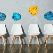 Depositphotos_202679476_L Vier weiße Stühle vor einer Wand mit bunten Papierknäueln, die eine Glühbirne symbolisieren.