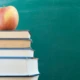 Ein roter Apfel liegt auf einem Stapel Bücher vor einer grünen Tafel.