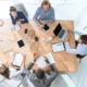 Eine Gruppe von fünf Personen sitzt an einem Tisch und arbeitet zusammen mit Notizbüchern, Tablets und Laptops.
