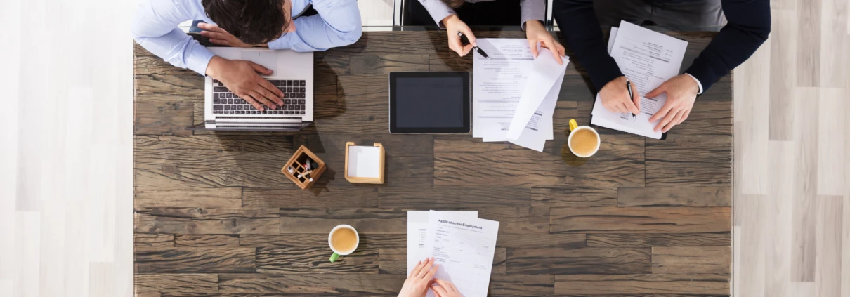 Vier Personen arbeiten an einem Tisch mit Laptops und Dokumenten. Kaffeetassen und Schreibutensilien sind verteilt.