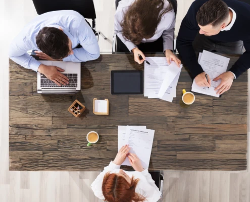 Vier Personen arbeiten an einem Tisch mit Laptops und Dokumenten. Kaffeetassen und Schreibutensilien sind verteilt.