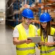 Zwei Personen in Schutzhelmen und Warnwesten stehen in einem Lager und schauen gemeinsam auf ein Tablet.