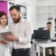 Zwei Kollegen in einem Büro betrachten gemeinsam ein Tablet, während im Hintergrund zwei weitere Personen an einem Schreibtisch arbeiten.