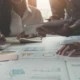 Mehrere Personen arbeiten an einem Tisch, der mit Finanzberichten und Diagrammen bedeckt ist, während sie mit einem Tablet und einem Taschenrechner arbeiten.