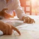 Zwei Personen arbeiten an einem Schreibtisch und diskutieren über Dokumente und Diagramme, die auf dem Tisch verteilt sind. Eine Person zeigt auf ein Papier, während die andere ihre Hand über ein Tablet hält.