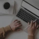 Das Bild zeigt eine Person, die an einem Laptop arbeitet. Auf dem Schreibtisch befinden sich eine Tasse Kaffee, ein Notizbuch, Bleistifte und ein Stifthalter. Die Person hält einen Stift in der rechten Hand und tippt mit der linken Hand auf der Tastatur.